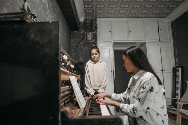 A student observes her teacher practicing piano in a cozy home music studio setting.