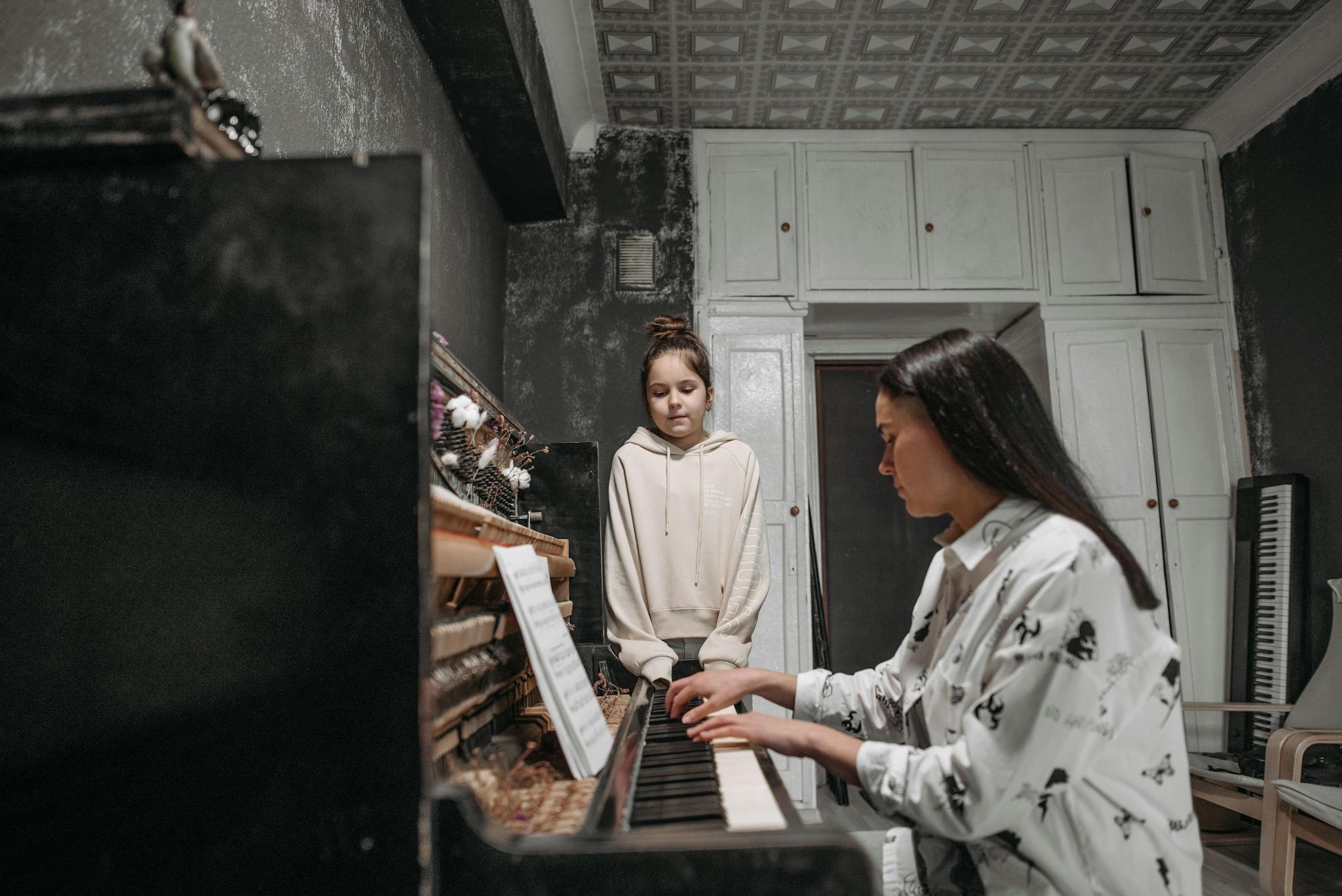 A student observes her teacher practicing piano in a cozy home music studio setting.
